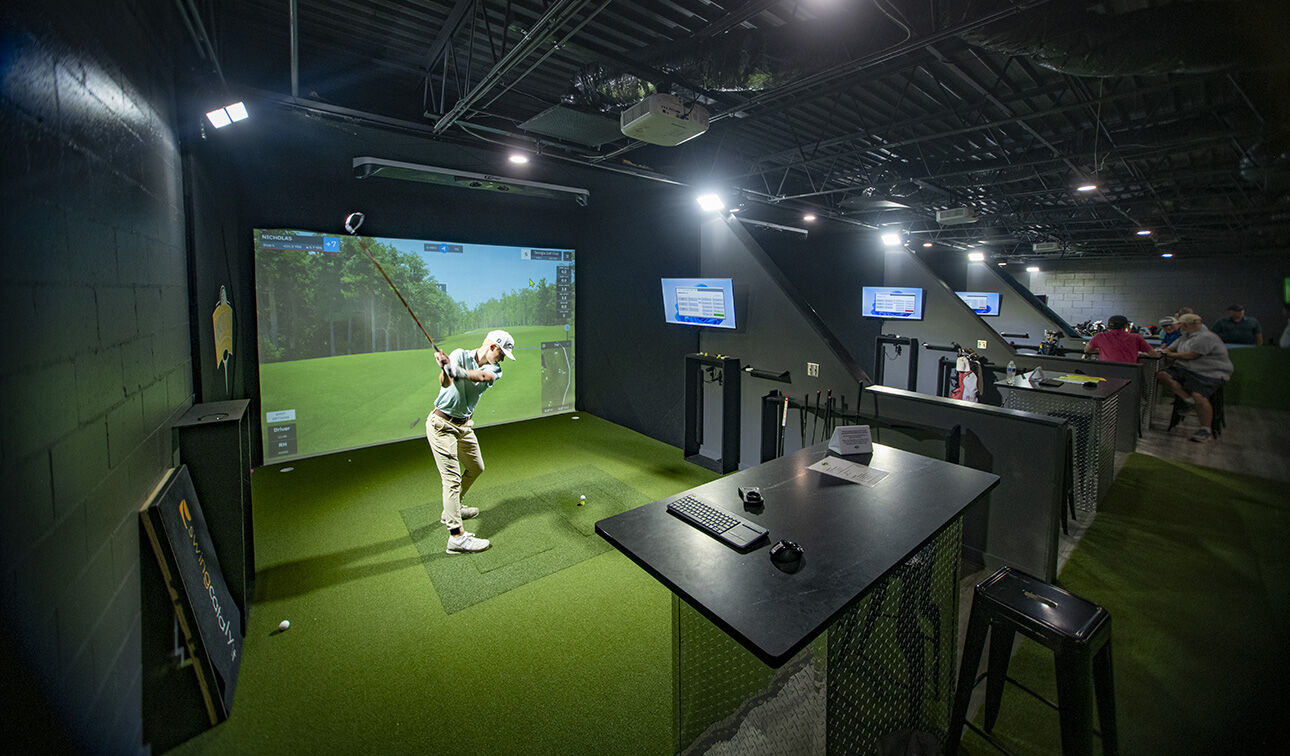 A player mid-swing in a hitting bay at Magic City Indoor Golf, facing the
                    lit simulator screen with the monitor station alongside