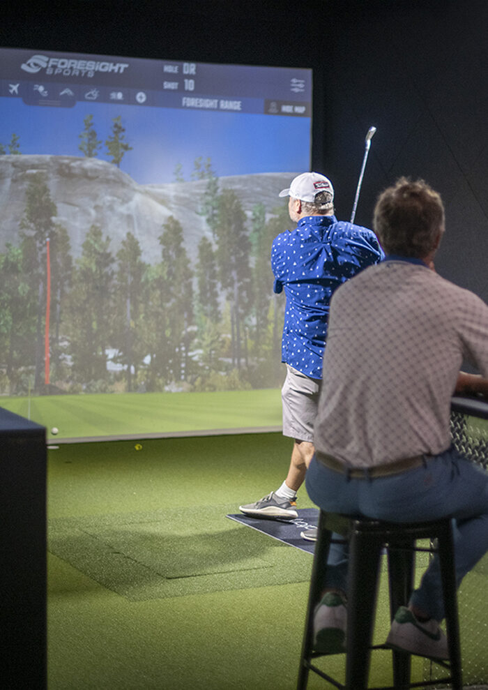 A PGA professional watching from a stool while a player hits into the
                    simulator screen, launch-monitor data displayed above
